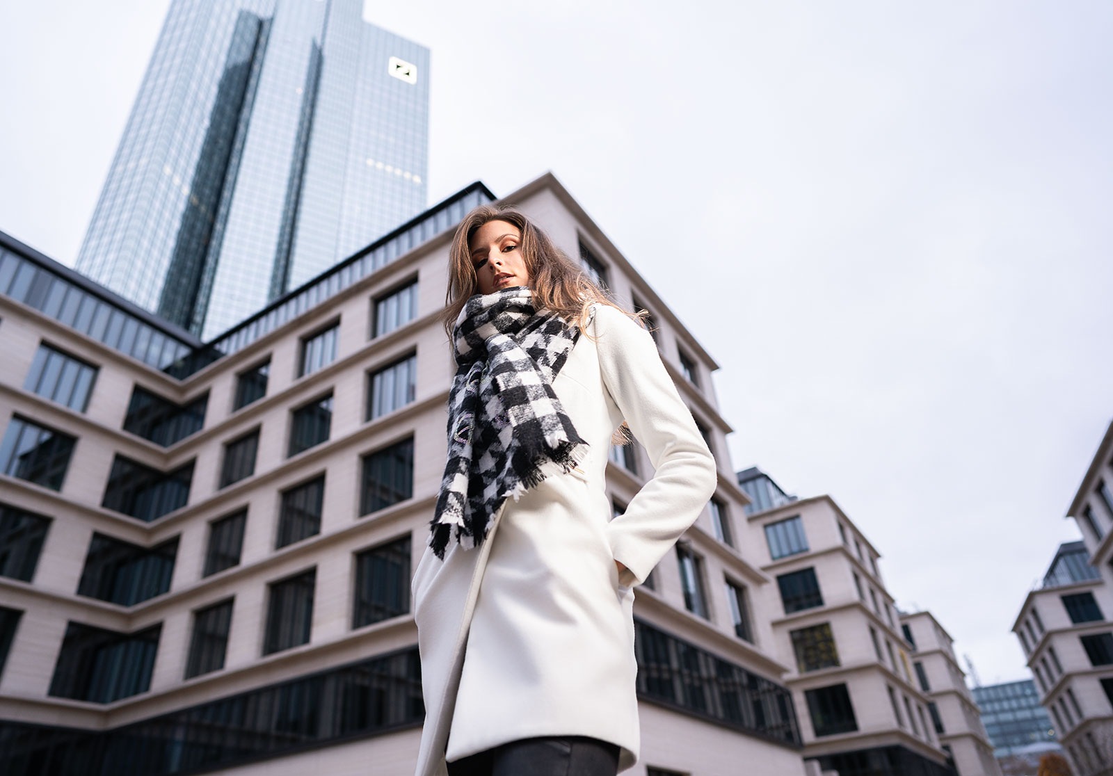 Fashion Editorial Portrait einer Frau in elegantem weißen Mantel und High Heels, fotografiert vor moderner Frankfurter Architektur. Cinematic Street-Style-Fotografie mit urbanem Flair, cleanem Licht und architektonischer Tiefe. Ideal für Modemagazine, Editorials und Kampagnen mit Fokus auf Stil, Selbstbewusstsein und minimalistisches Design.